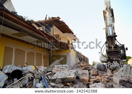 Construction/demolition site with debris pile in foreground, excavator and partly destroyed building in background.