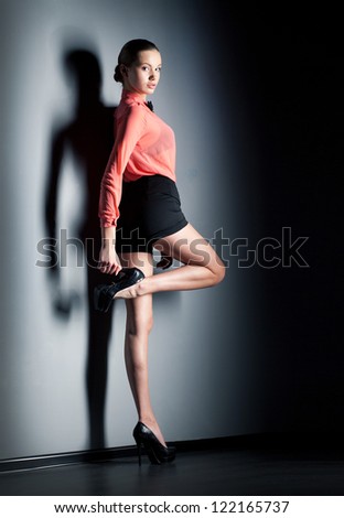 graseful brunette model in a blouse and shorts in full growth and her hand on the heel. model posing on a gray wall, illuminated hard studio lighting