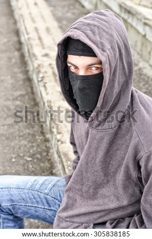 unrecognizable young man wearing black balaclava sitting on old stairs, looking at the camera