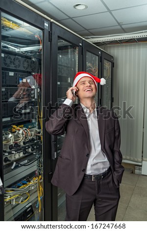 Network Engineer laughs in the server in Santa Claus hat with mobile phone