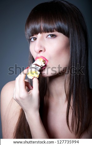 Sexy brunette female model with long straight healthy hair, glossy red lips, holding tasty donut with her hands, eating it with great pleasure and temptation, looking at camera. Black background