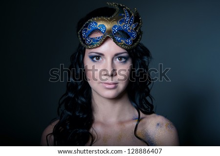 Pretty brunette girl with shiny healthy curly long hair, shoulders naked, wearing elegant makeup, colorful ornate golden blue traditional mask on forehead, looking at camera. Black background