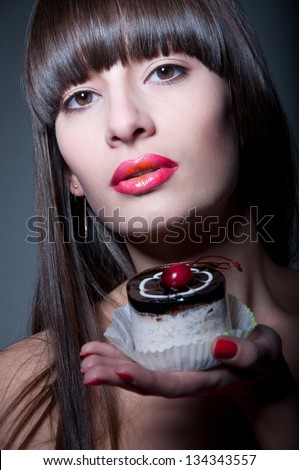 Pretty brunette woman model with long straight hair, glossy red lips, holding and offering tasty tart cake with cherry on her hand, looking at camera. Dark gray background, copy space