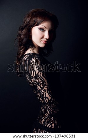 Profile portrait of beautiful young woman model with long hair in locks and curls wearing black transparent lace dress, looking at camera. Black background