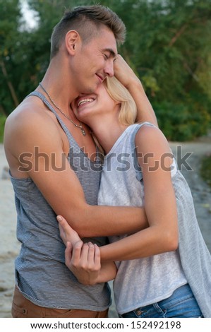 Young pretty couple of man and woman wearing tees and jeans, being in love close to each other, embracing, smiling, kissing, feeling happy together. At sunset