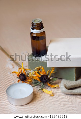 Spa products natural white and green bar of soap on olive oil on a small towel, aroma oil bottle for aroma therapy, two dry yellow flowers near soap, sea salt, candle on the wooden table surface