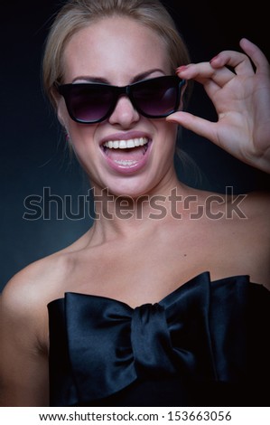 Funny young blond woman model wearing stylish black top with big silk bow, sun glasses, bright pink lipstick, having fun, laughing, shouting, smiling with toothy smile at camera. Black background