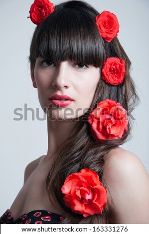 Pretty young woman model with red lips, elegant long brunette hair weaved into wave with big fresh terracotta roses, black corset underwear with red lips pattern. Looking at camera