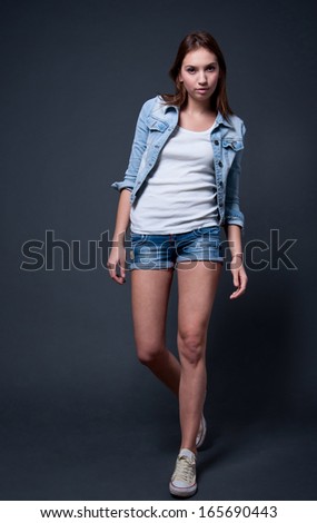 Cute stylish pretty young girl model with very long legs wearing white top, blue denim shorts, blue cotton jacket, sneakers, walking, looking at camera. In studio over gray background