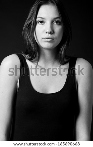 Black and white studio portrait of young pretty model woman with dishveled hair style, wearing black tee top, looking at camera with surpsire and hope. Over black background
