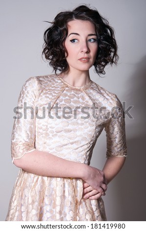 Studio portrait on gray of beautiful brunette woman model with curly hair, tender makeup wearing light golden dress with narrow belt, looking at camera