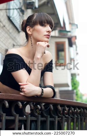 Pretty slim young brunette woman model wearing black off-the-shoulder top, elegant earrings, beautiful hairstyle, leaning over balustrade of a balcony, looking somewhere, dreaming and enjoying the day