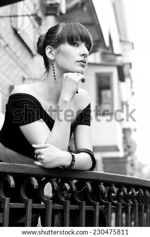 Black white portrait of pretty slim young woman model in black off-the-shoulder top, elegant earrings, nice hairstyle, leaning over balustrade of balcony, looking somewhere, dreaming, enjoying the day