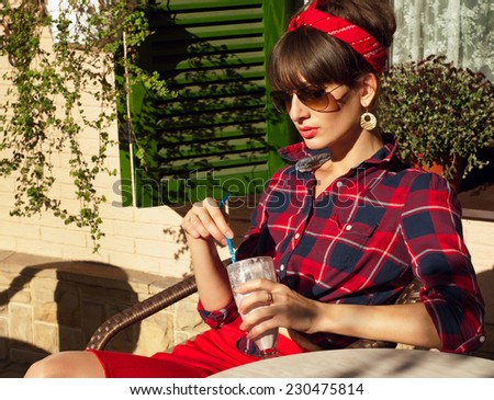 Pretty brunette woman wearing red skirt, checked shirt, band, aviator sunglasses, resting in a wickered chair in a street cafe, drinking her cold milk shake cocktail. On a hot summer day