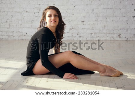 Beautiful elegant gymnastics athlete girl wearing long black dress, sitting on the floor, resting. Over white brick wall background  in studio