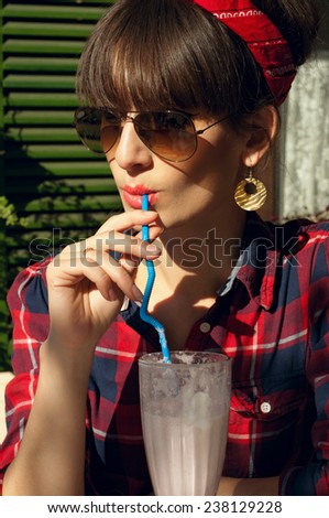 Pretty brunette woman wearing red skirt, checked shirt, band, aviator sunglasses, resting in a street cafe, drinking her cold milk shake cocktail. On a hot summer day