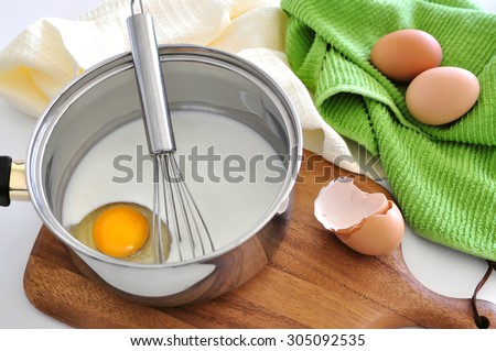 Cooking dough for pancakes: whisking milk with eggs in a pan on a wooden board, shell of broken eggs aside, several kitchen towels and napkins at background, copy space