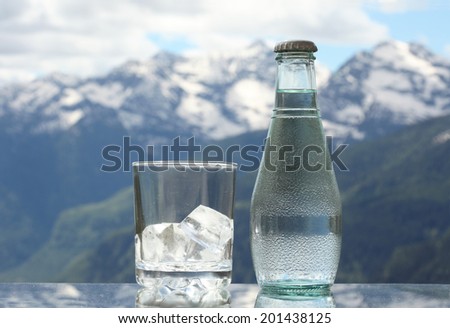 Mineral water bottle and glass with ice cubes, Alps mountains on the background
