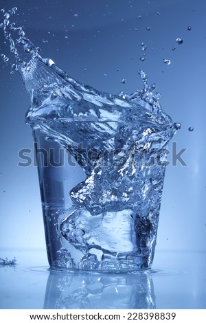 Water glass with a splash of falling ice cube isolated on blue background