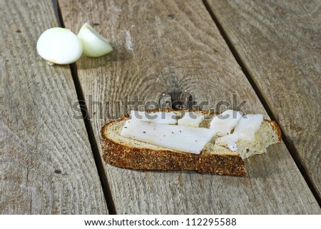 Bitten piece of soft bread with white lard is on a rough wooden table. Cutting in half onion bulb is blur on background.