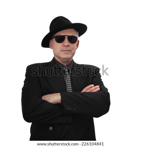 Mafia is immortal - dangerous looking middle aged man in classic black suit and hat with sunglasses stands with his arms crossed isolated on white background