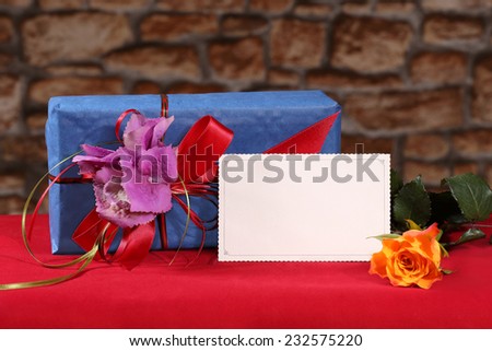 Empty postcard stands in front  of decorative holiday gift box together with yellow rose flower on red table on blurred stone all background