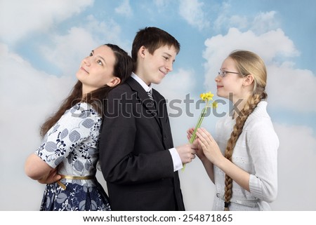 Teenage boy stands back to back with one girl and gives flowers to another on cloudy blue sky background - Inconstancy and volatility of youth concept