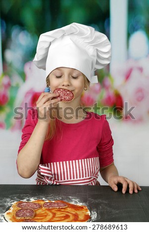 Cute little girl in chief hat enjoys smell of sausage when preparing pizza - cooking, food and pleasure concept