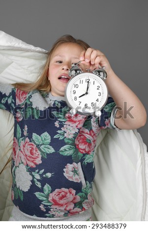 Little girl with blanket and alarm clock in hand shows she woke up too late on gray background - Morning time and lateness concept