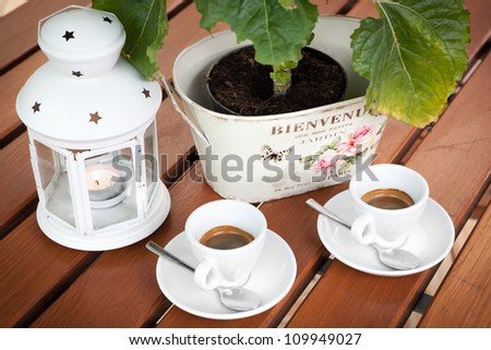 Two cups of espresso coffee on the table in the garden cafeteria restaurant. Decorative lamp and green plants in the background. White cups with silver spoons.