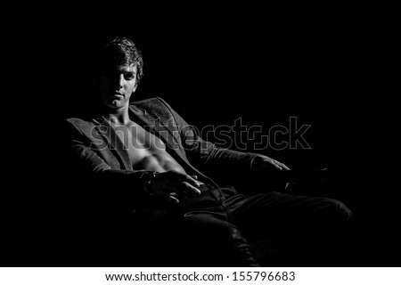 Young handsome macho man with muscle abdominal and open jacket sitting in armchair. Black and white studio shoot