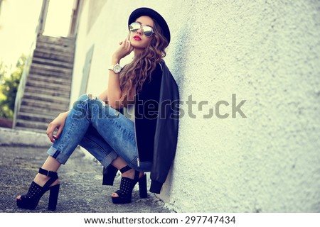 fashion model with long curly hair wearing sunglasses sitting and posing outdoor. Jeans, shoes, hat, jacket.