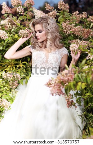 Beautiful bride posing outdoor among the flowers. Wedding dress, curly blond hair.