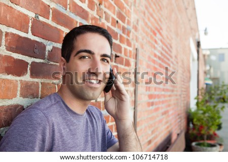 A cute guy on a mobile phone outdoors by a brick wall, looking at camera with a smile.