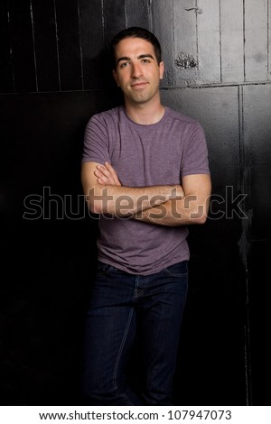 Good looking guy leaning on a black wall with arms crossed wearing a purple shirt and jeans