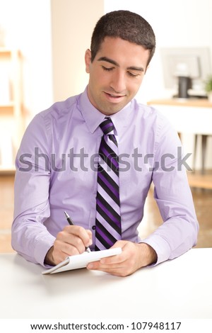 Handsome guy looking down writing on pad of paper with pen wearing a purple business shirt and tie.