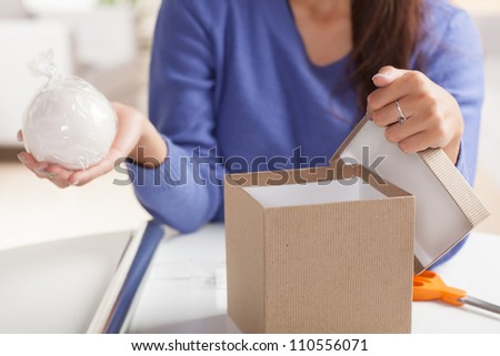 Adorable Hispanic woman wrapping gift with silver wrapping paper and brown box  in living room at table wearing blue shirt.