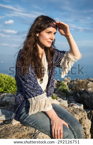 Portrait of  woman looking forward, sitting and relaxing on a natural coastal rock high up, contemplating the sea against a blue sky. Well being healthy lifestyle.. Outdoors beauty.