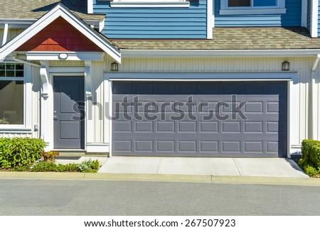 Family townhouse on sunny day, British Columbia, Canada. Double garage with concrete driveway and asphalt road in front. Residential townhouse with entrance door beside wide garage door.