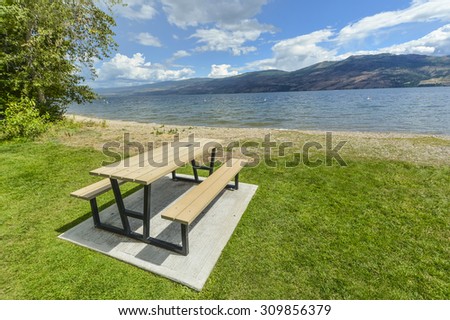 Picnic table and benches near Okanagan lake. Table and benches at rest area close to waterfront with mountains and cloudy blue sky background.