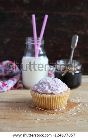 Cupcake with blackcurrant jam and coconut and cream cheese frosting in paper linens and a jar of jam and milk bottle with straws on a wooden surface