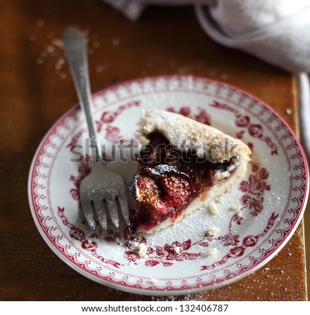 Piece of fig and raspberry galette pie with icing sugar on a dessert plate with fork and napkin
