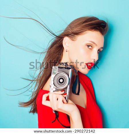 Portrait of a beautiful young brunette woman in a red dress in the studio on a blue background with a camera in his hands