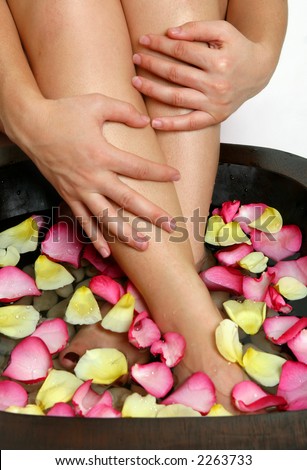 A woman luxuriates her weary feet with a relaxing footsoak.  Pink and yellow flower petals float on the warm water releasing a heavenly scent.
