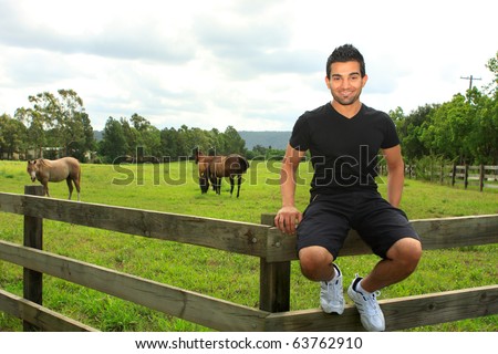 A smiling happy young man sitting on a wooden post and rail fence of a field with horses grazing.