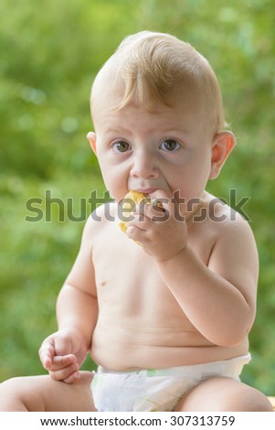 Cute naked baby eating bread sitting on grass, summer day