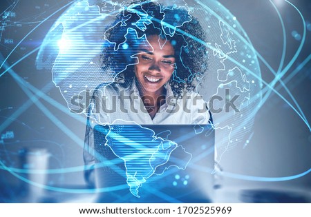 Cheerful young African American woman using laptop in blurry office with double exposure of futuristic network interface and planet hologram. Concept of internet and communication. Toned image