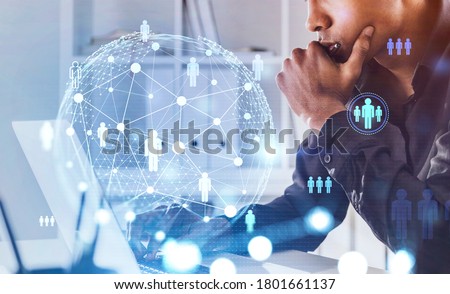 Side view of serious young African American businessman using laptop in blurry office with double exposure of social network interface. Concept of HR. Toned image
