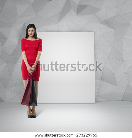 Beautiful brunette woman in a red dress is holding fancy shopping bags. Contemporary wall and an empty whiteboard on the background.