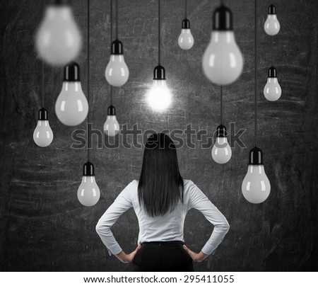 Rear view of the brunette lady who is looking at the hanging light bulbs. a concept of searching new ideas. Black chalk board background.